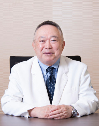 理事長兼学長の竹之下誠一氏が白衣を着て微笑む姿。背景は淡い色合いの壁。