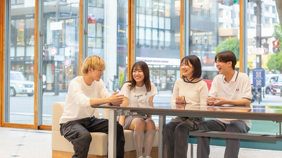 大きな窓の前のテーブルに座り、談笑している四人の若者たち。明るい自然光が差し込むカフェのような室内。