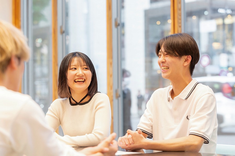 室内で4人の若者がテーブルを囲んで談笑している。背景には大きな窓から外の風景が見える。