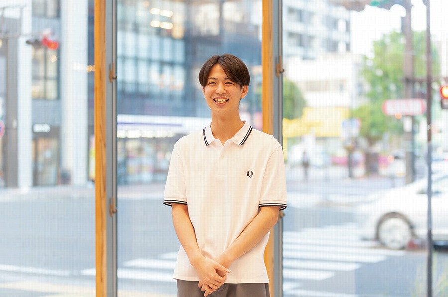 明るい窓辺で白いシャツを着た男性が笑顔で立っている。背景には街の風景が広がっている。