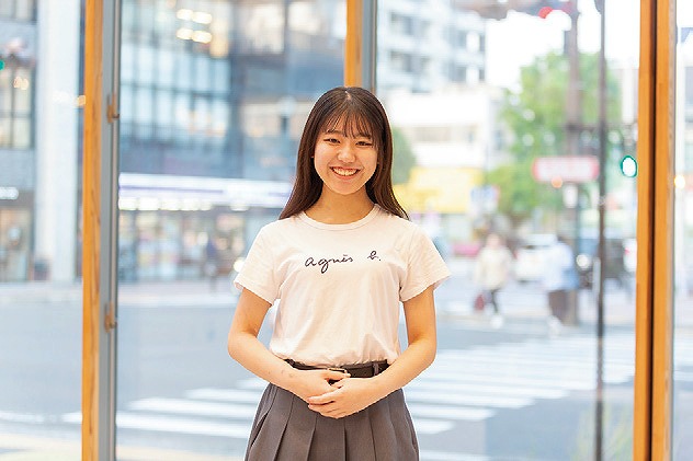 交差点を背景に白いシャツを着た女性が微笑んで立っている。明るい日差しが差し込み、街中の窓辺での一瞬。