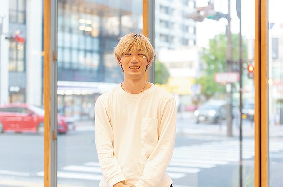 大きな窓から見える街並みを背景に、カジュアルな白いシャツを着た若い男性が微笑んで立っています。
