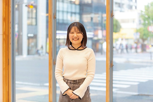 明るい窓辺に立ち微笑んでいる女性。背景には街並みが広がり、交差点の横断歩道が見える。