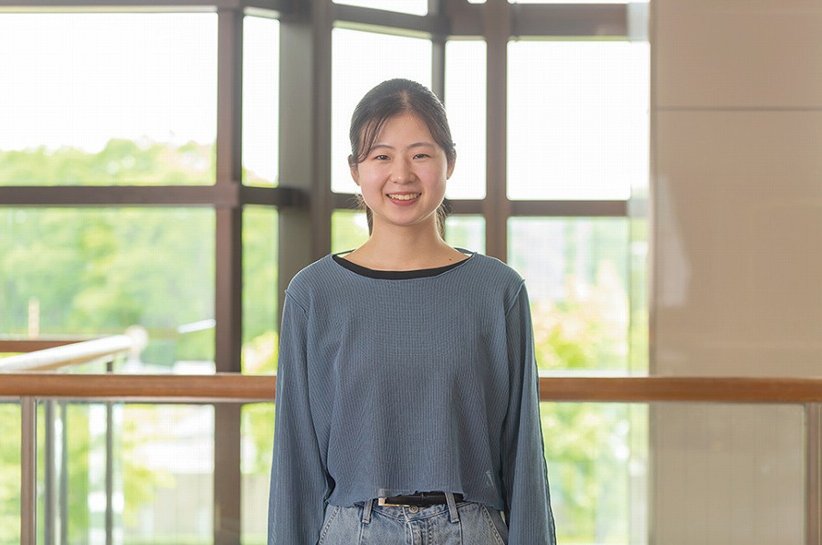 ガラス窓を背景に微笑む若い女性。カジュアルな服装で、明るい室内に立ちリラックスした様子を見せている。