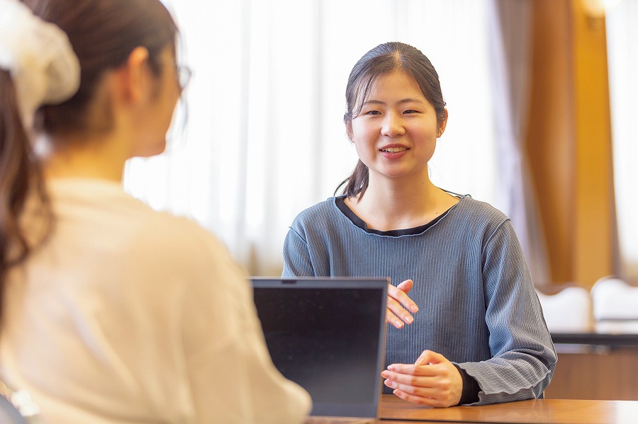 机で対面して話す二人の女性。前景の女性は背を向け、後景の女性が微笑みながら説明している様子。