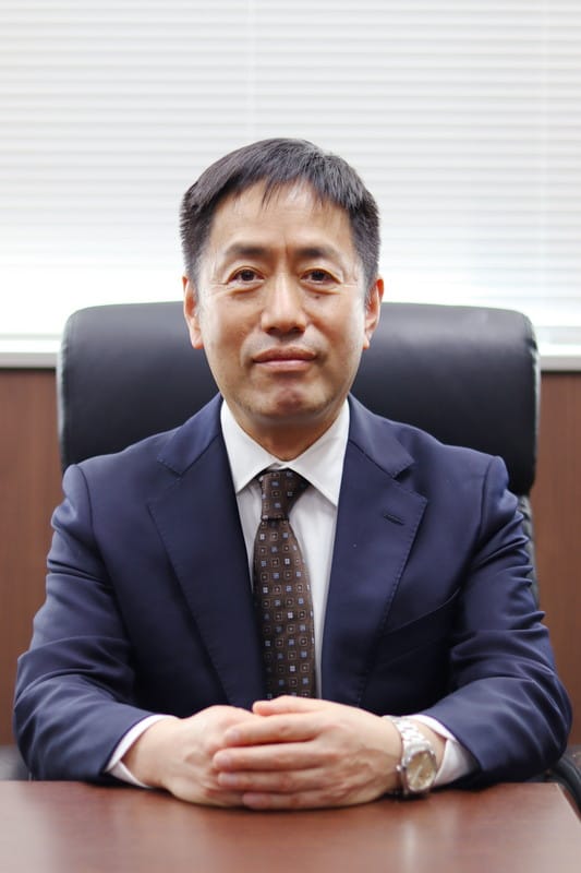 大学院保健科学研究科長の林博史氏が椅子に座り、真剣な表情でカメラを見つめているポートレート。