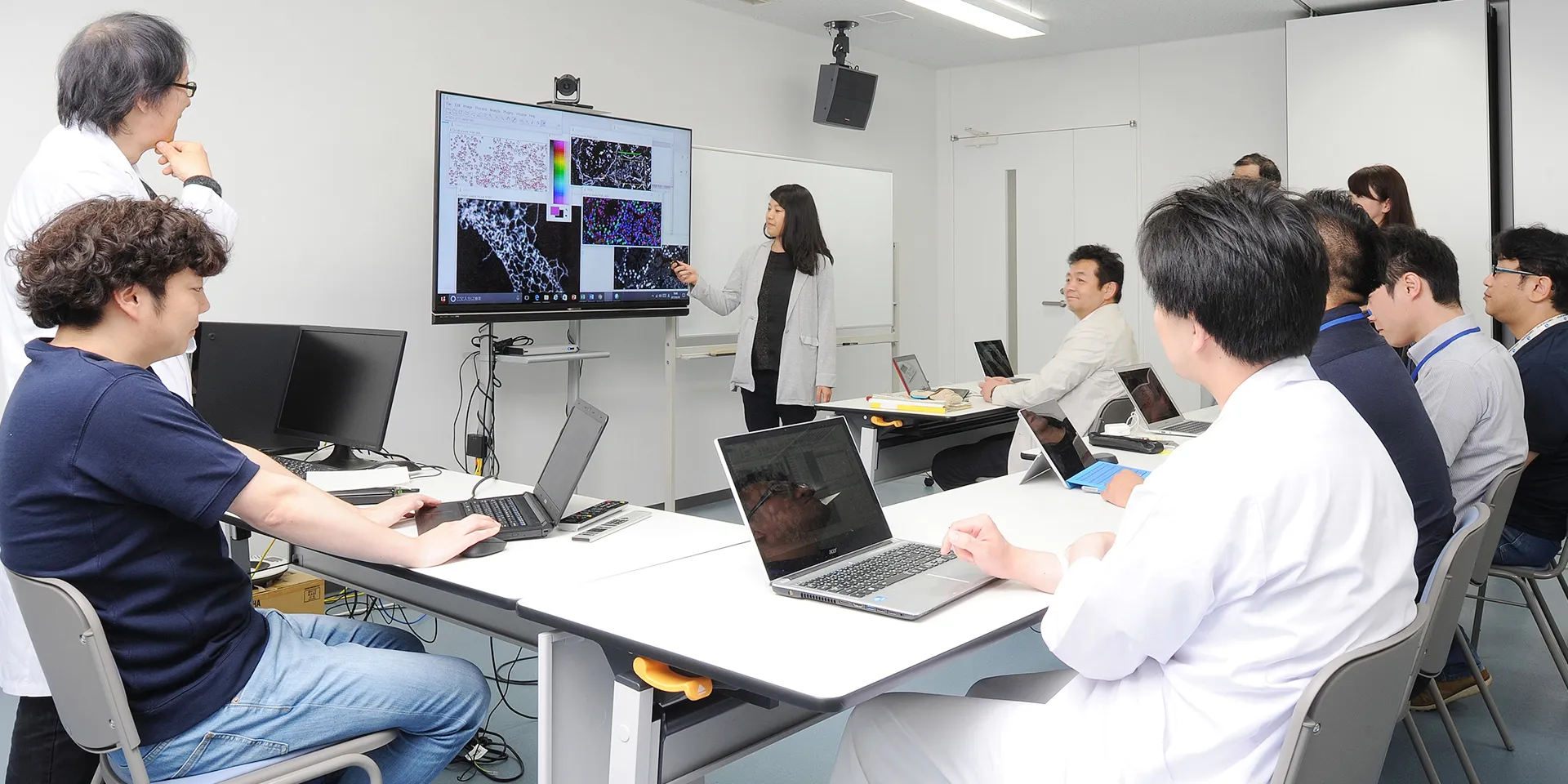 大学院の研究室で、プレゼンテーションを行う女性とそれを聴く研究者たちの様子。モニターには顕微鏡画像が表示されている。