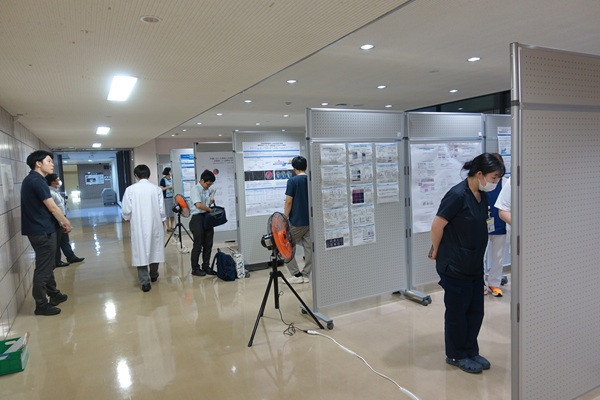 大学内で学生や専門家がポスターを使ってディスカッションする様子。背景には窓があり、外の光が差し込んでいる。
