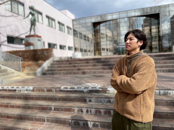 福島県立医科大学の前で考え込む男性、背景には大学の建物と像が見える。