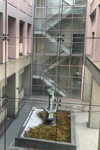 福島県立医科大学の中庭にある彫刻と緑地、背後にはガラス張りの階段が見える風景。