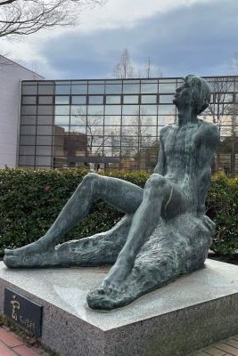 福島県立医科大学のキャンパスにある、座っている人物のブロンズ像。背景にはガラス張りの建物が見える。