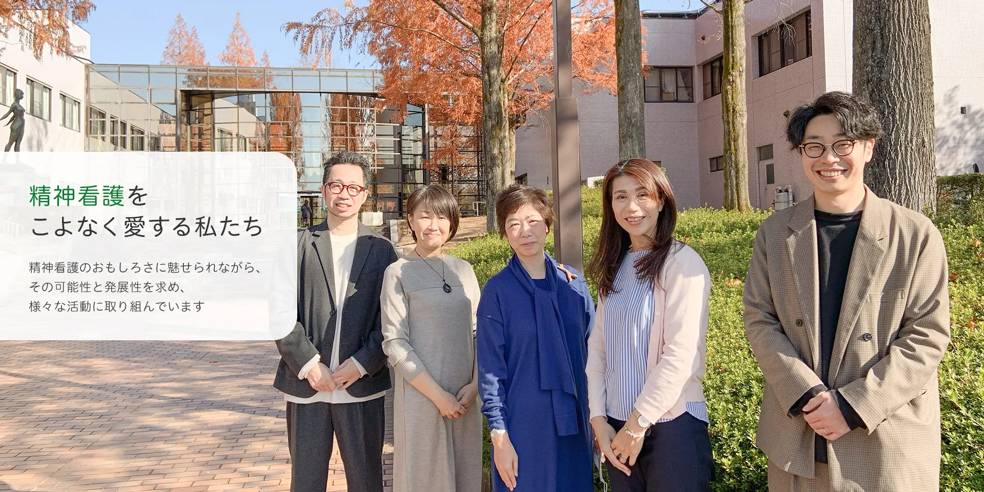 キャンパス前で並んで微笑む5人の人々。バックにはガラス張りの建物と秋の木々が見える。