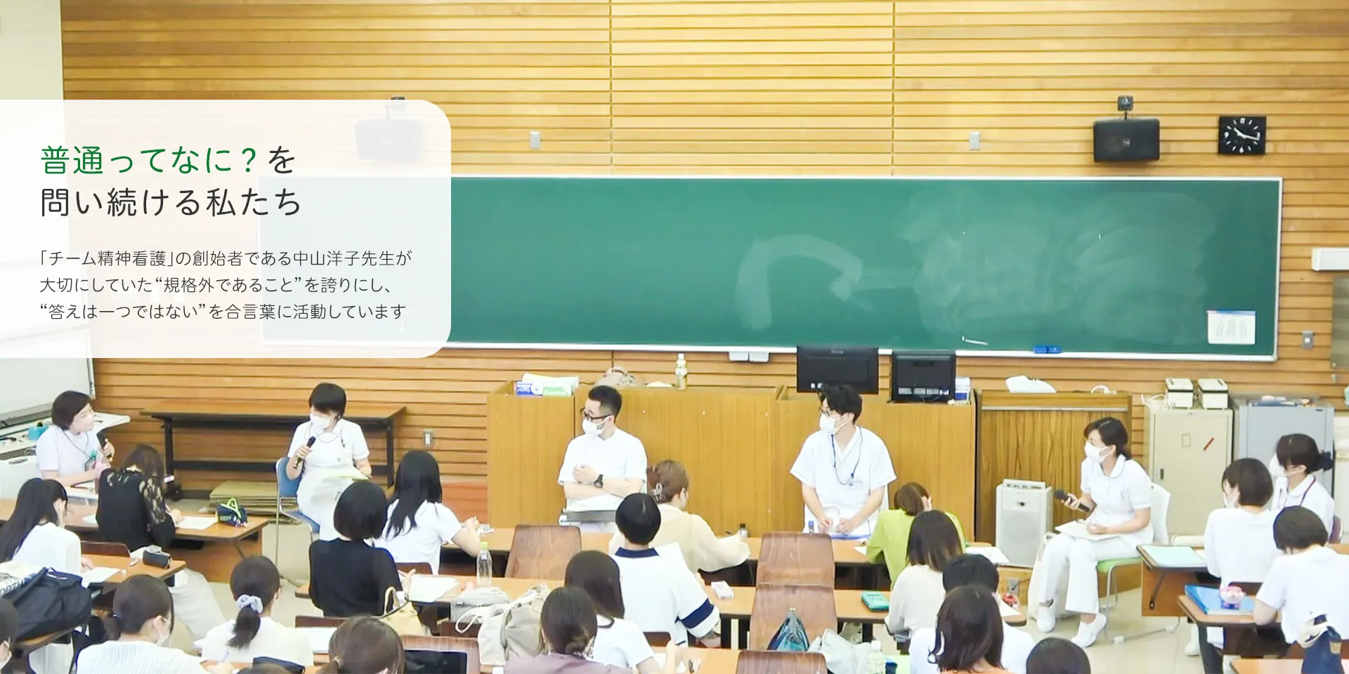 教室で授業を受けている学生たち。前方で数名の講師がマイクを持ちながら話をしている。