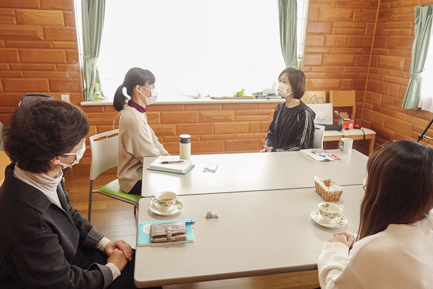 マスクを着用した4人の人々が室内でテーブルを囲み談話中。各自の前にノートや飲み物が置かれている。