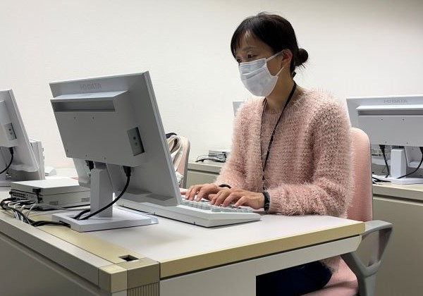パソコンの前で作業をする女性がマスクを着用し、真剣な表情でキーボードを打っている様子。