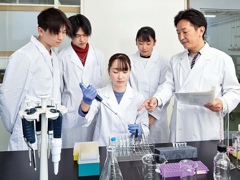 白衣を着た学生と指導者が実験室でピペットを使って実験を行っている様子。周囲には試験管や実験器具が並ぶ。