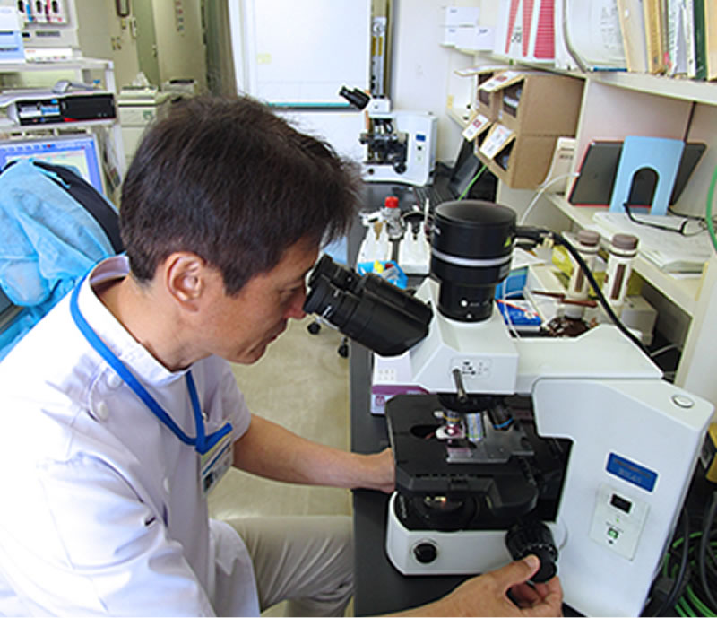 白衣を着た男性が研究室で顕微鏡を使ってサンプルを観察している様子。背景には他の研究機器や書籍が並んでいる。