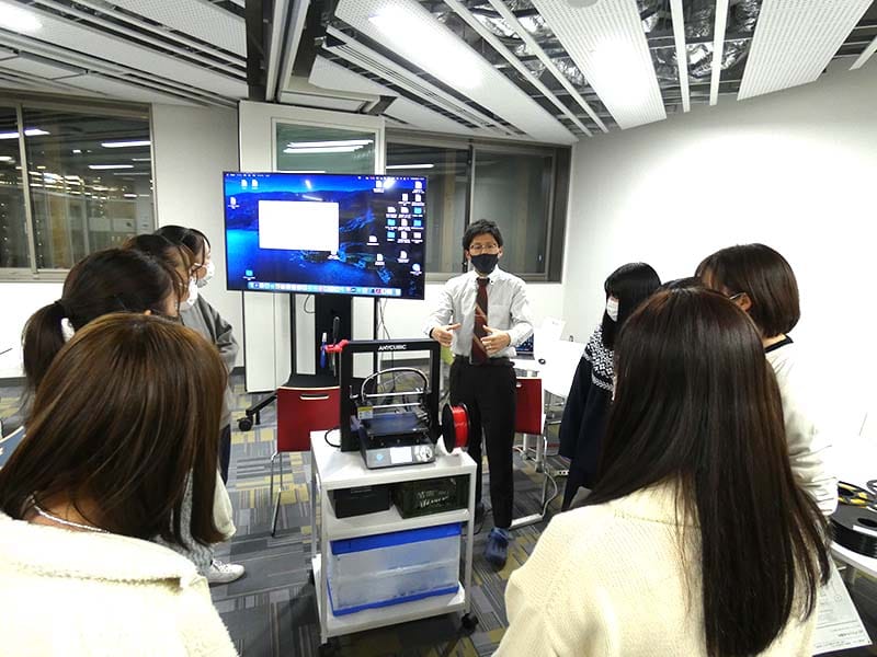 講師が学生たちに3Dプリンターの使い方を説明している教室の様子。背景には大きなモニターが映っている。