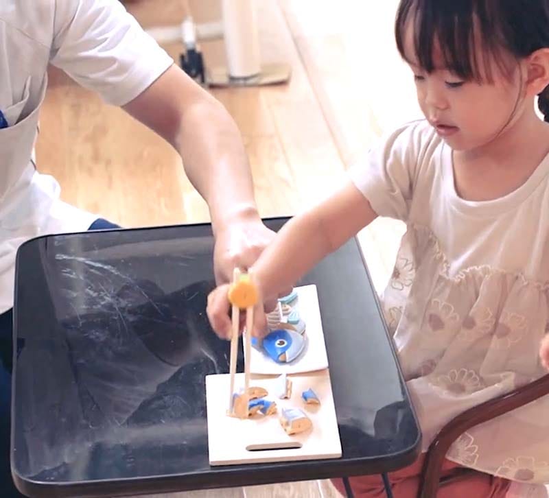 小さな女の子が積み木を使ってテーブルで遊んでおり、隣に大人がサポートしている。