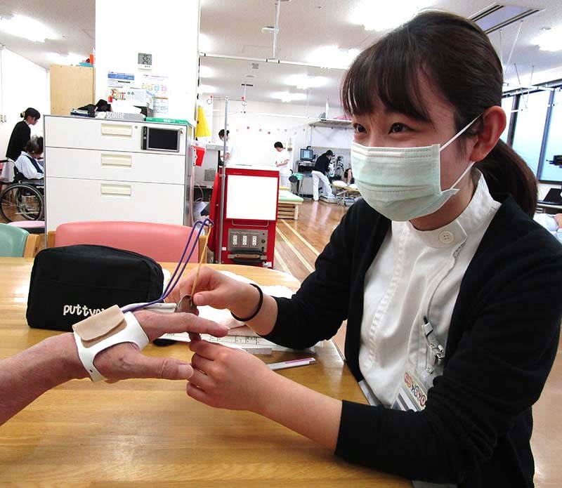 マスクをした看護師が、患者の手に装置を装着してサポートしている診療室の様子。背景に他の医療スタッフがいる。