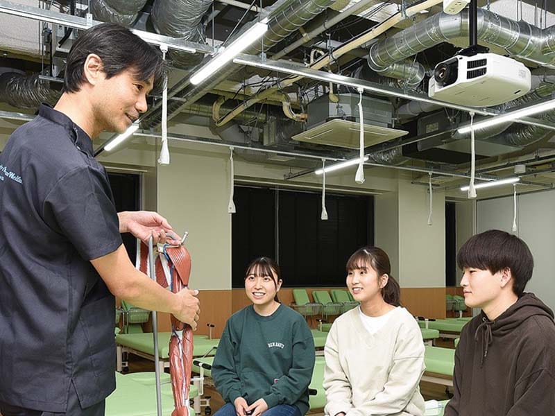 医療教室で、講師が模型を使って学生に説明している様子。学生たちは座って講師を見ている。背景にはプロジェクターと天井設備。