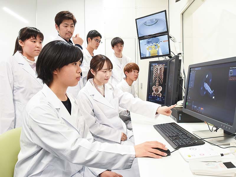 白衣を着た学生たちがパソコンを囲んで医学的な画像を見ている様子。講師らしき人物が指示を出している。