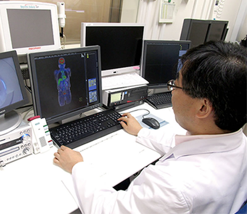 白衣を着た人物が、複数のモニターに囲まれた医療用コンピュータの前で画像を確認している。