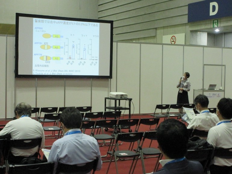 2021 BioJapanでの口頭発表の様子。スライドを使って研究内容を説明する講演者と、聴衆が座っている会場の風景。