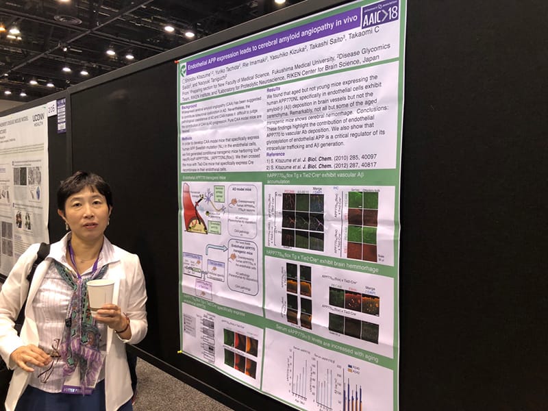 臨床検査学科の研究者がポスターの前で説明している様子。ポスターには神経変性疾患に関する研究内容が記載されている。