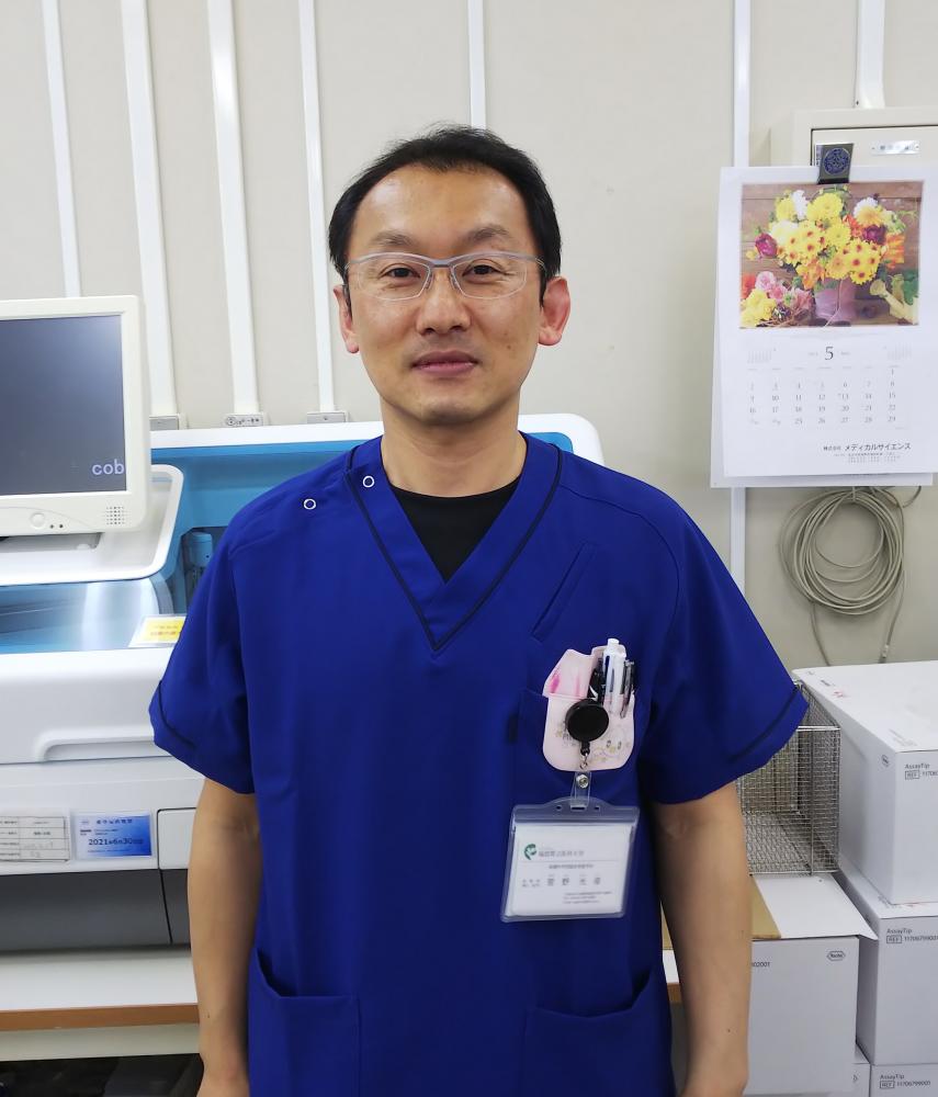 臨床検査技師の菅野光俊さんが青い作業着を着て微笑んでいる写真。背景には医療機器やカレンダーが見える。