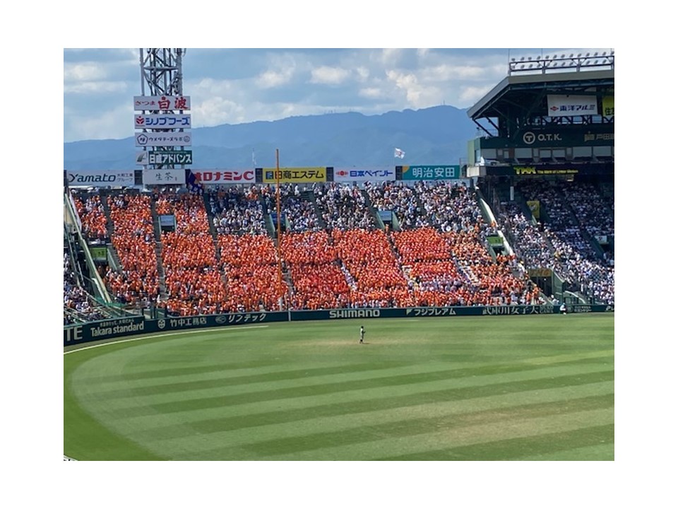高校野球の試合で、オレンジのユニフォームを着た大応援団が熱心に応援している様子。背景には青空と山々が広がる。