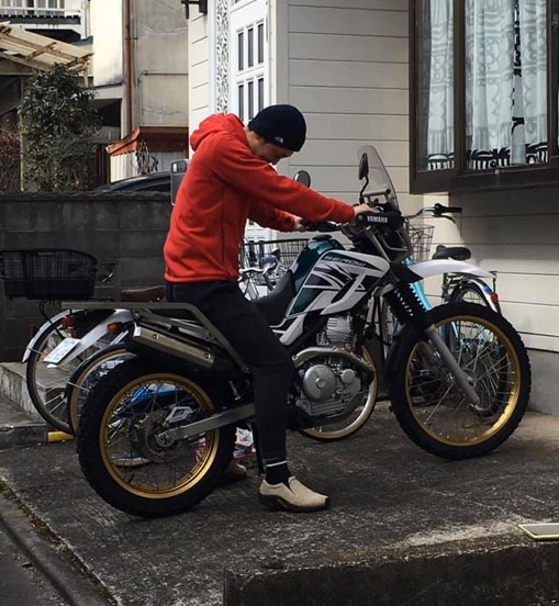 バイクに乗る男性が赤いパーカーを着ている様子。背景には他のバイクや建物が見える。