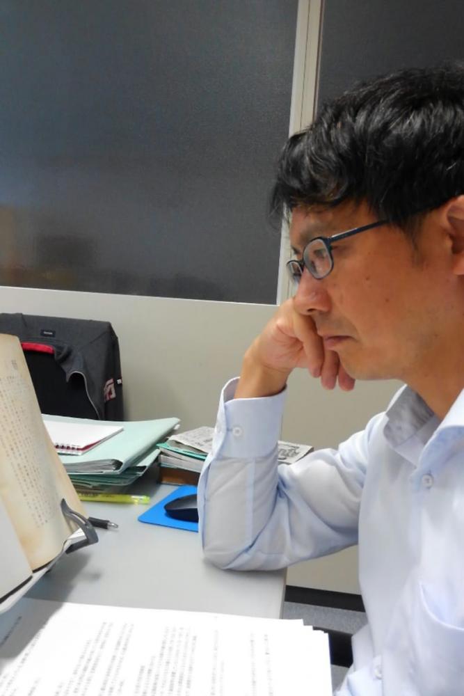 メガネをかけた男性が机に向かい、真剣に本を読んでいる様子。周囲には書類やノートが散らばっている。