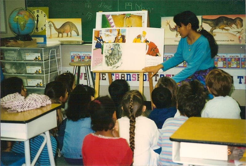 子供たちに絵本を読み聞かせる女性教師と、その様子を見守る生徒たちの風景。