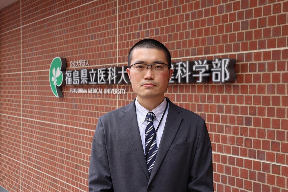 福島県立医科大学の看板の前に立つ男性の写真。スーツを着ており、真剣な表情をしています。