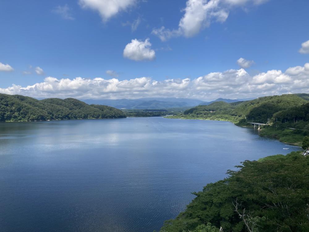 美しい青空と緑に囲まれた湖の風景。穏やかな水面が広がり、遠くには山々が見える。