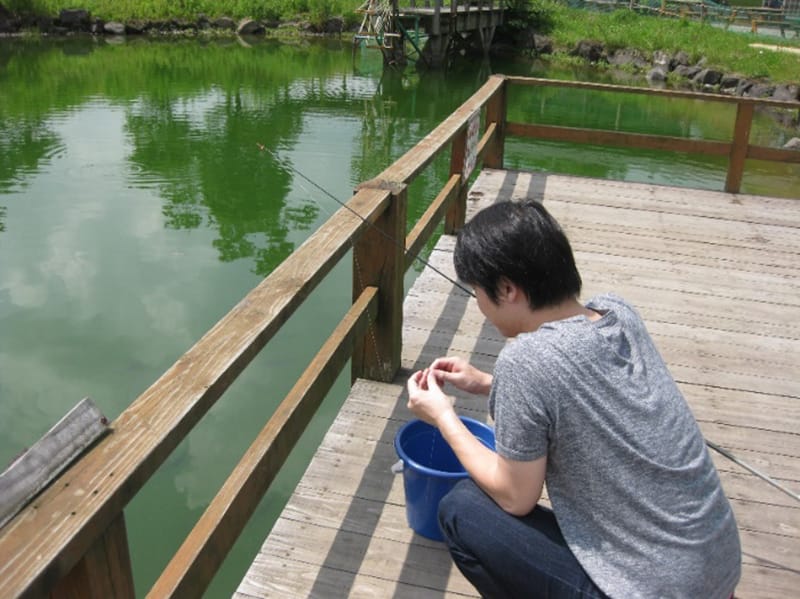 湖のほとりで青いバケツを持ちながら何かをしている人物の後姿。周囲には緑色の水面と木製のデッキが広がっている。