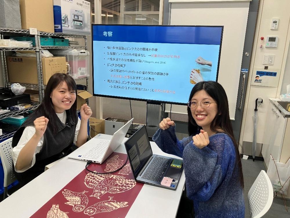 卒業研究に取り組む学生2名が、パソコンの前で発表準備をしている様子。背景には研究内容が表示されたモニターがある。