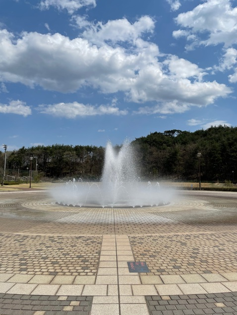福島県立医科大学の広場にある噴水と青空の風景。周囲には緑が広がり、穏やかな日差しが差し込んでいる。