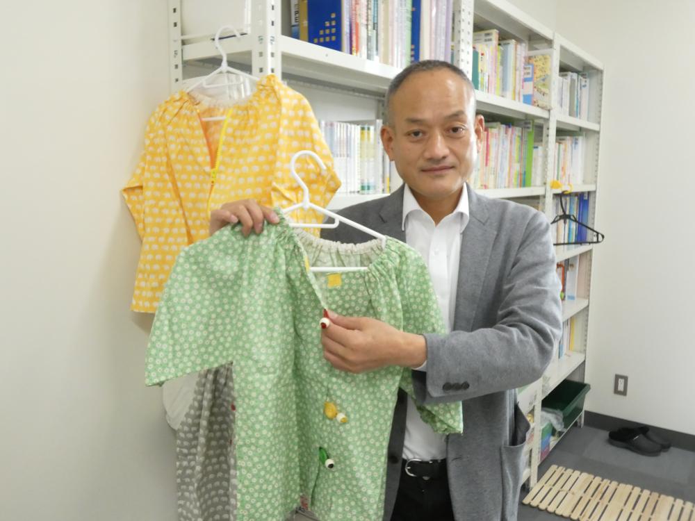 作業療法士が緑の服を持ちながら、背景に本棚と他の衣服が見える室内の様子。
