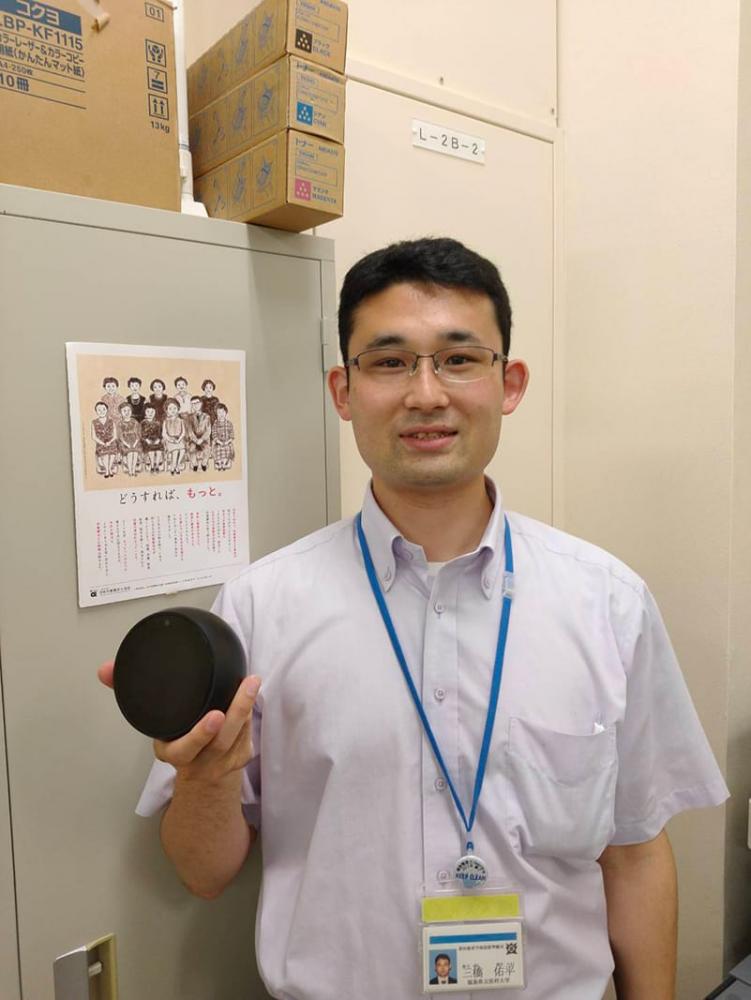 作業療法士が黒い円形の物体を持ちながら微笑んでいる写真。背景には掲示物や段ボールが見える。
