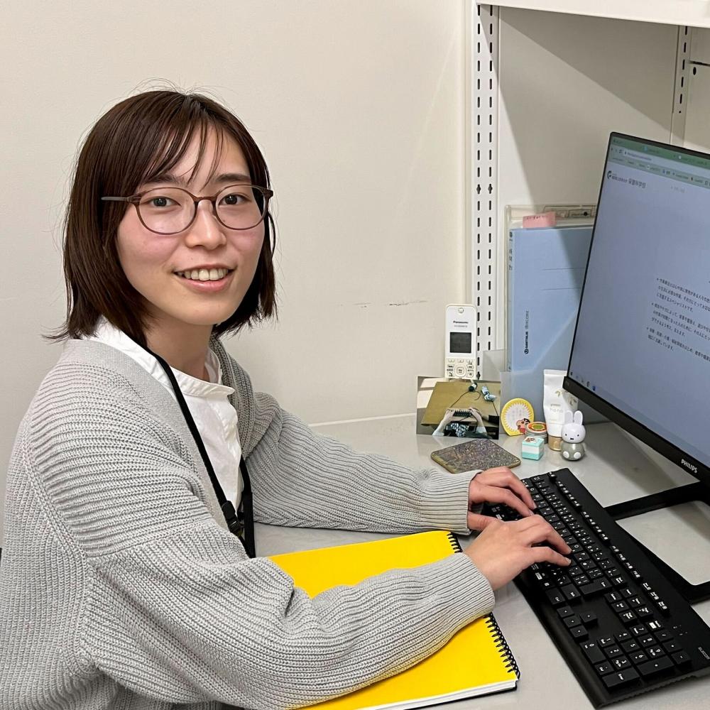デスクでパソコンを操作しながら微笑む女性の写真。彼女はグレーのセーターを着ており、黄色いノートが目の前に置かれている。