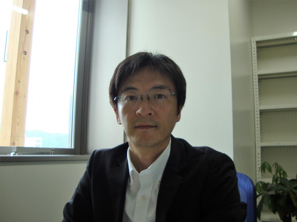 作業療法士としての自己紹介を行う男性が、オフィスでカメラに向かって微笑んでいる様子。