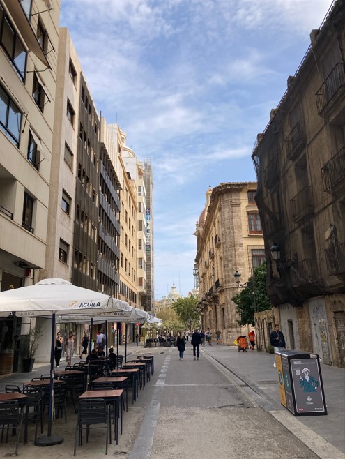バレンシアの市街地の通り、カフェのテラス席が並び、賑やかな人々が行き交う風景。