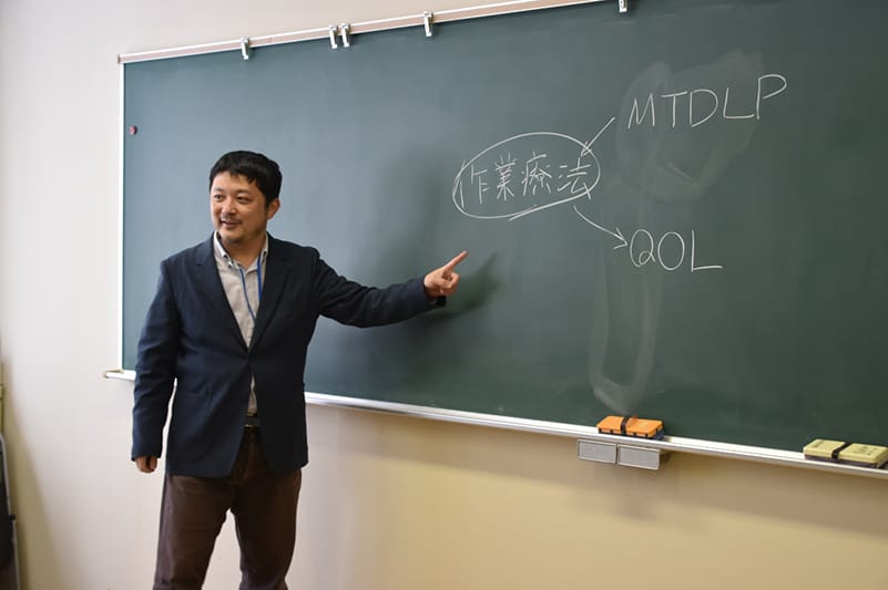 講義中の講師が黒板を指し示し、「作業療法」や関連用語を説明している様子。