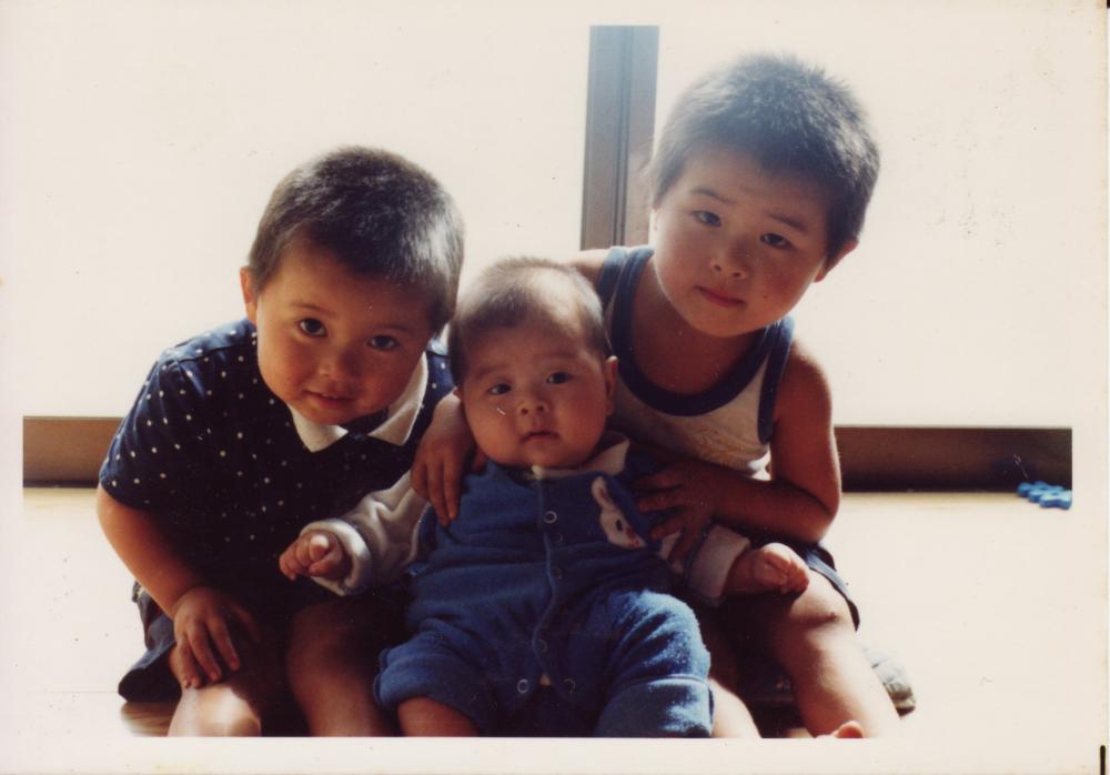 三人の子供が仲良く座っている写真。左側の子供は水玉模様のシャツ、中央の赤ちゃんは青いオーバーオール、右側の子供はタンクトップを着ています。