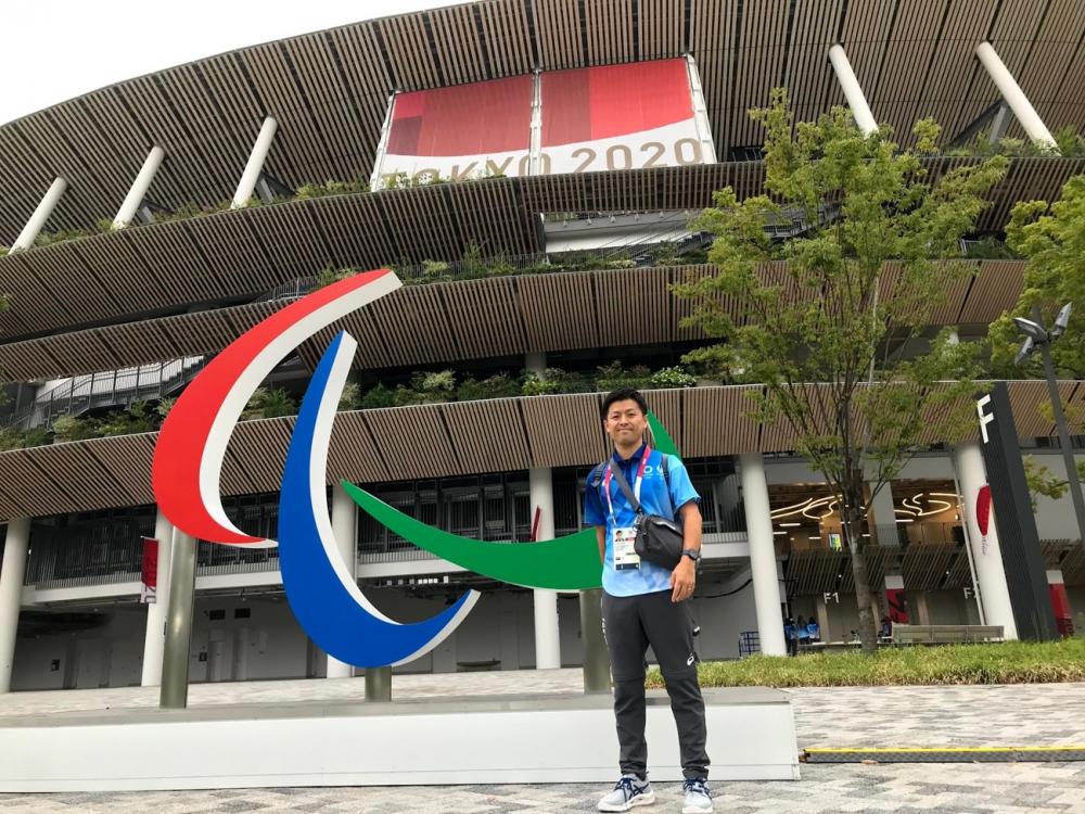 東京2020パラリンピックの会場前で、笑顔の男性が立っている様子。背景にはスタジアムが見える。