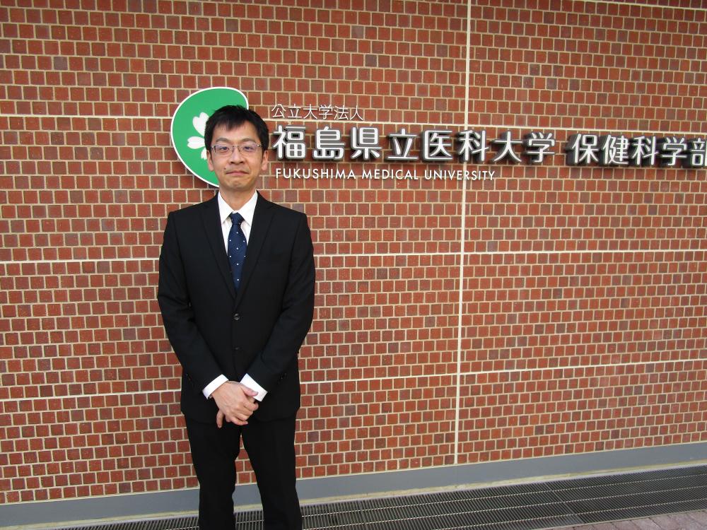 福島県立医科大学の看板の前に立つ男性がスーツを着ている写真。