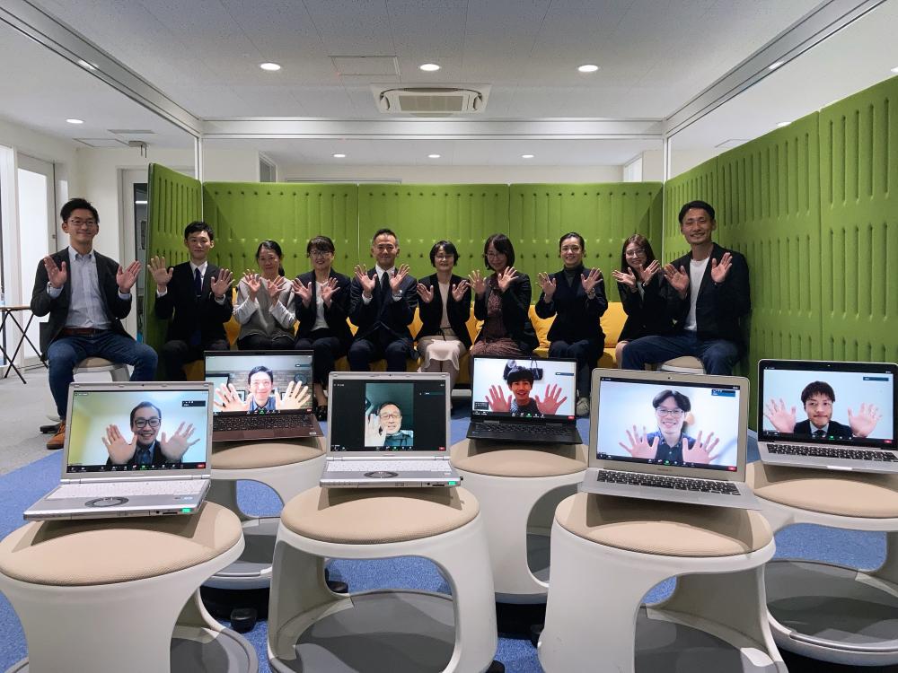 学術大会の運営に参加するメンバーが、オンライン参加者と共に手を挙げている様子。背景には緑のパネルが配置されている。
