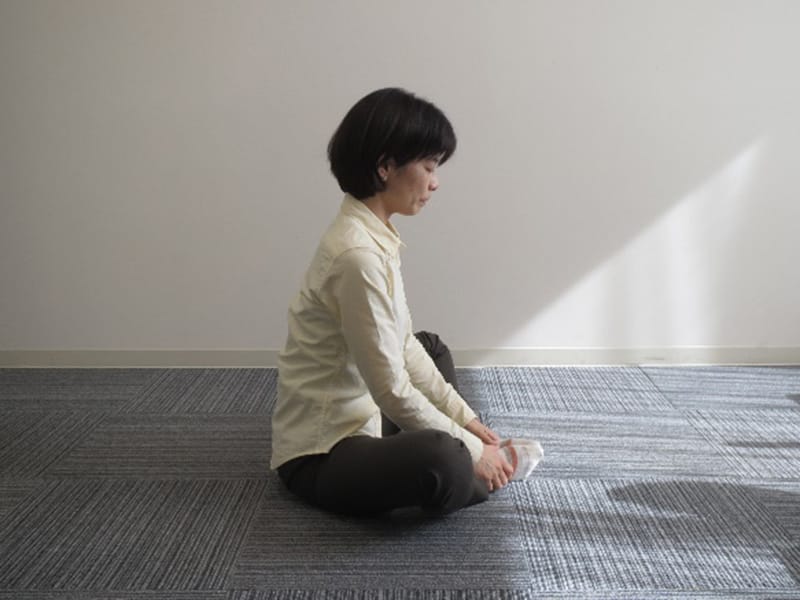座ってストレッチをする女性の姿。明るい光が差し込む室内で、リラックスした雰囲気。